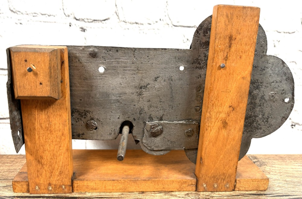 A 17th-century French wrought iron twin spring bolt door lock featuring detailed foliate scroll engravings and lobed metal plates, mounted on a light timber display stand. View V