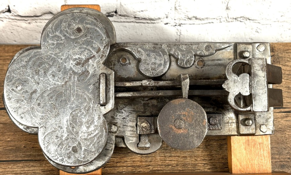 A 17th-century French wrought iron twin spring bolt door lock featuring detailed foliate scroll engravings and lobed metal plates, mounted on a light timber display stand. View VI