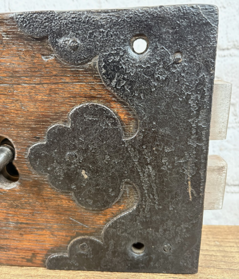 A 19th-century rectangular oak cased stock lock with decorative black iron corner plates, a central keyhole, and an inserted iron key. View V