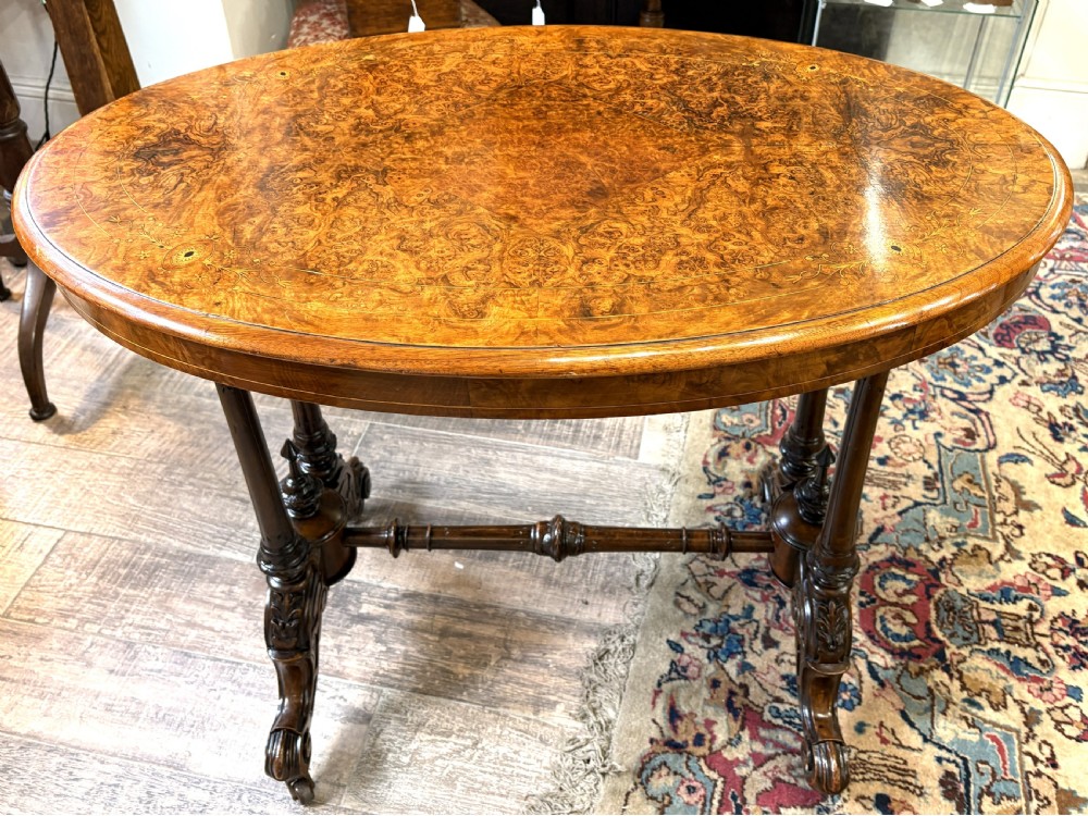 An oval Victorian burr walnut stretcher table featuring marquetry inlays, turned twin-column supports, a central stretcher rail, and carved legs with castors.