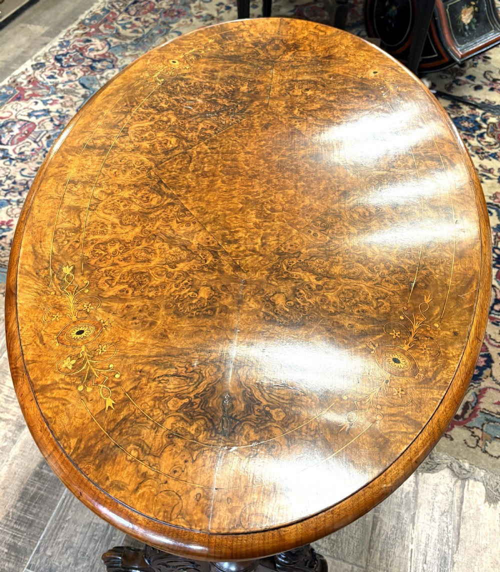 An oval Victorian burr walnut stretcher table featuring marquetry inlays, turned twin-column supports, a central stretcher rail, and carved legs with castors. View II