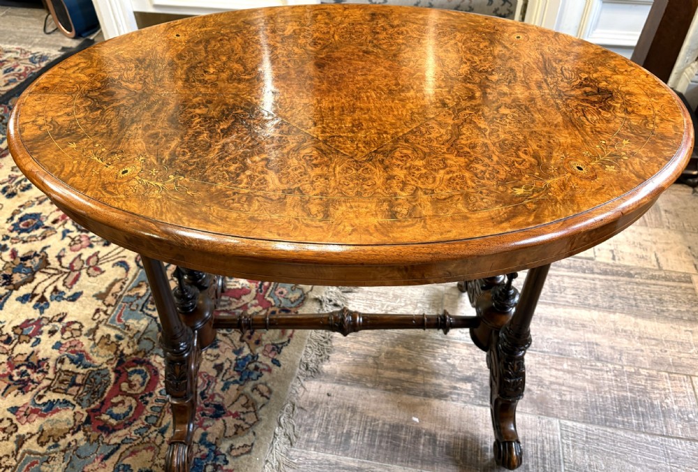 An oval Victorian burr walnut stretcher table featuring marquetry inlays, turned twin-column supports, a central stretcher rail, and carved legs with castors. View III