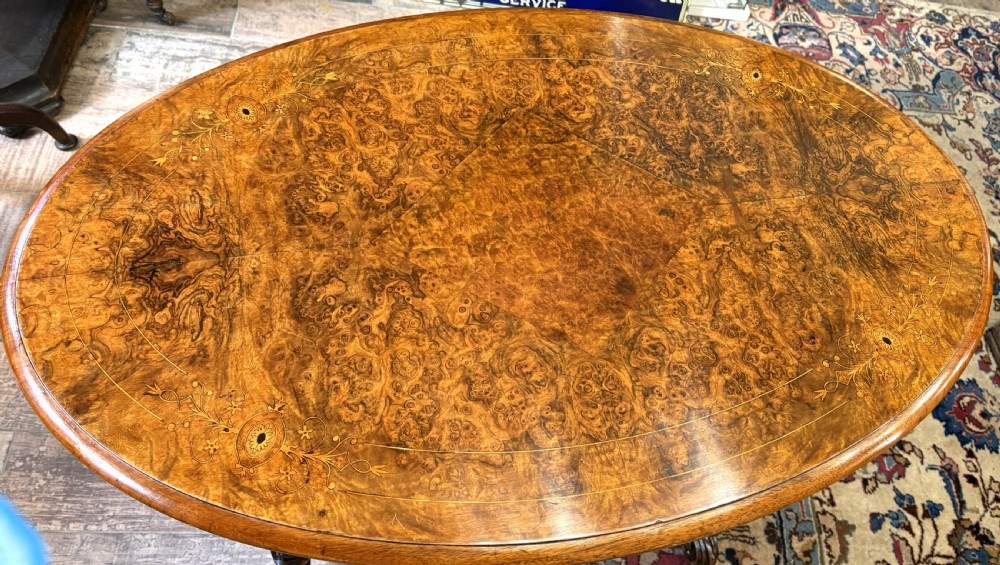 An oval Victorian burr walnut stretcher table featuring marquetry inlays, turned twin-column supports, a central stretcher rail, and carved legs with castors. View IV