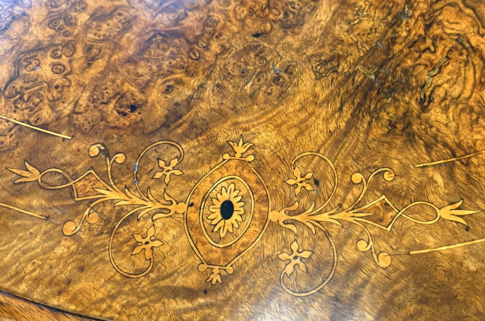 An oval Victorian burr walnut stretcher table featuring marquetry inlays, turned twin-column supports, a central stretcher rail, and carved legs with castors. View V