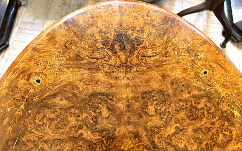 An oval Victorian burr walnut stretcher table featuring marquetry inlays, turned twin-column supports, a central stretcher rail, and carved legs with castors. View VI