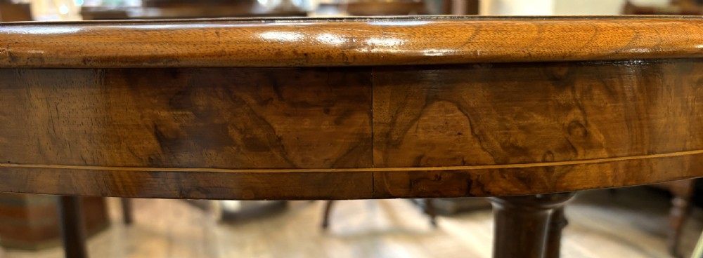 An oval Victorian burr walnut stretcher table featuring marquetry inlays, turned twin-column supports, a central stretcher rail, and carved legs with castors. View X