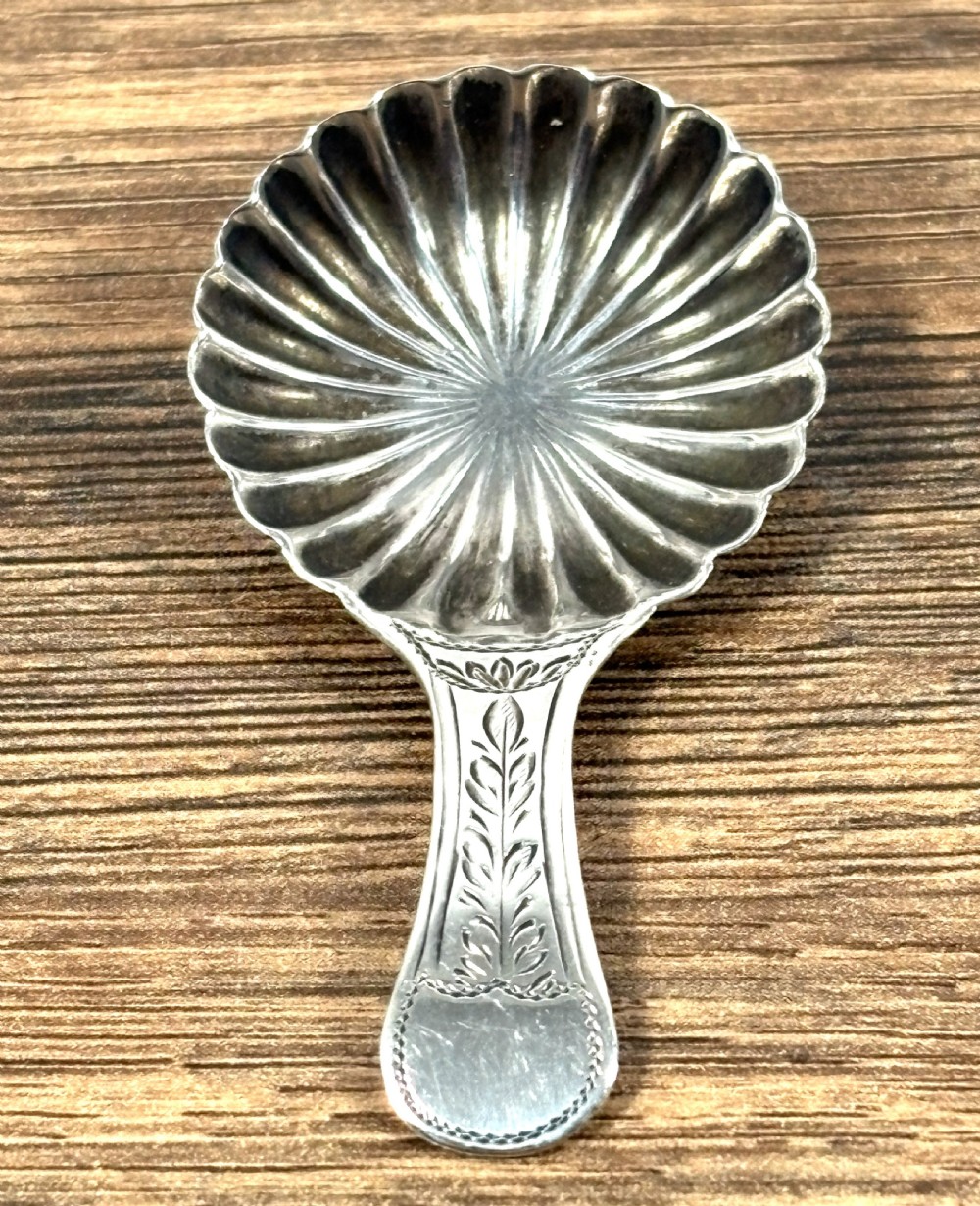 A Georgian sterling silver caddy spoon featuring a fluted shell-shaped bowl and a short handle with engraved leaf detail.