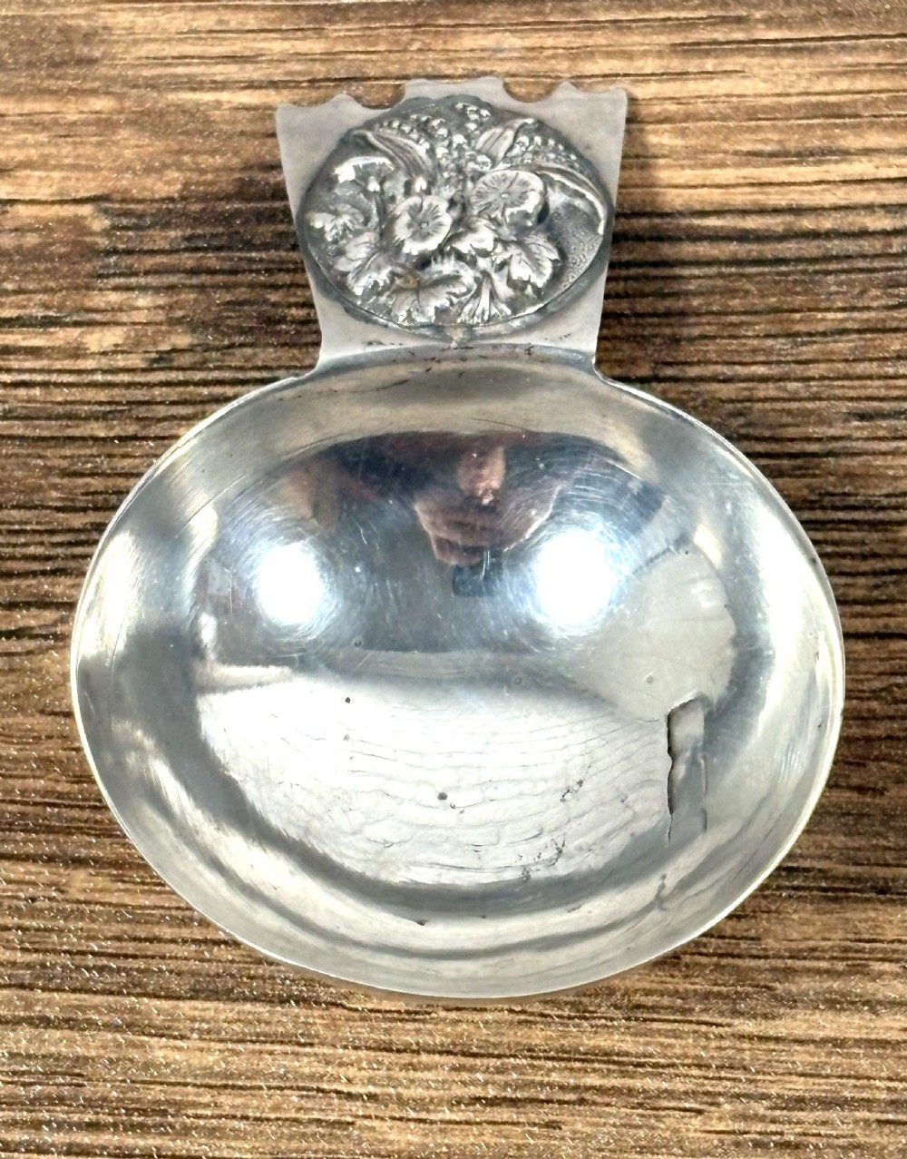 An Edwardian sterling silver caddy spoon featuring a smooth circular bowl and a short flat handle decorated with embossed floral detail.