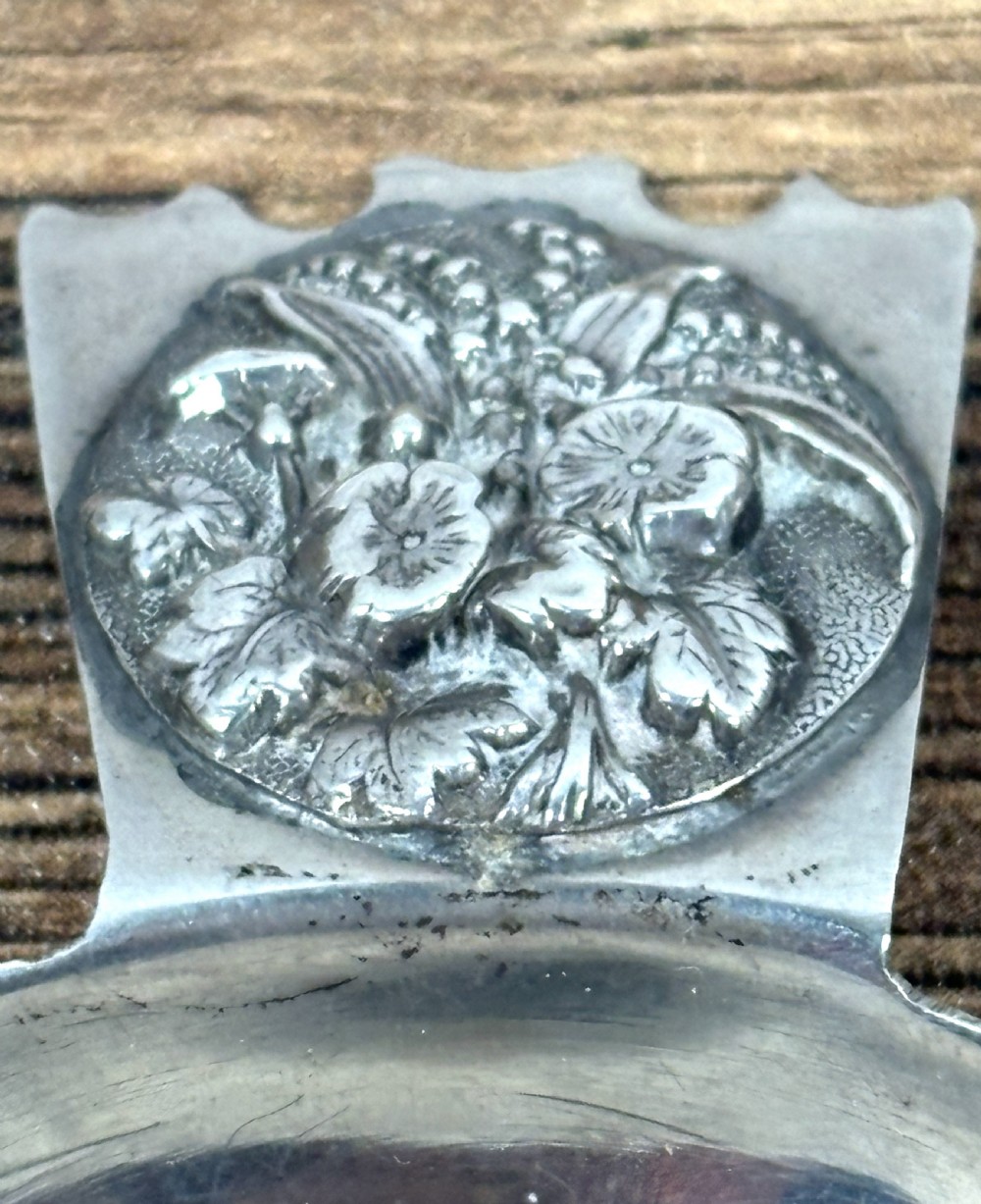 An Edwardian sterling silver caddy spoon featuring a smooth circular bowl and a short flat handle decorated with embossed floral detail. View III