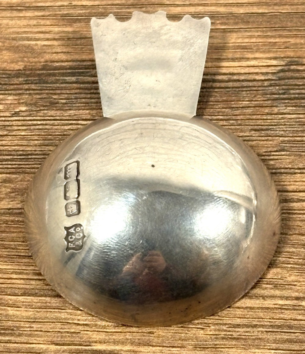 An Edwardian sterling silver caddy spoon featuring a smooth circular bowl and a short flat handle decorated with embossed floral detail. View IV