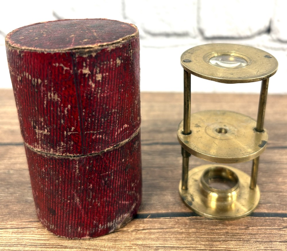 A Georgian brass travelling botanical microscope featuring three circular stages alongside its original red textured leather cylindrical case.