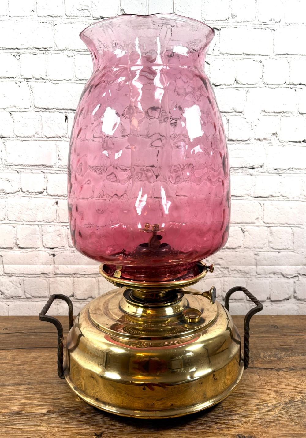 An antique Victorian oil lamp featuring a stepped polished brass base with twin wrought iron handles and a large dimpled cranberry glass hurricane shade.