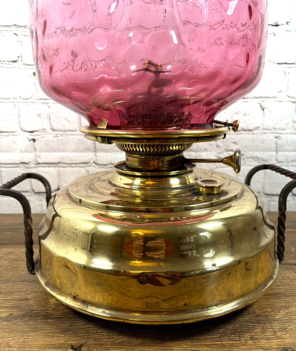 An antique Victorian oil lamp featuring a stepped polished brass base with twin wrought iron handles and a large dimpled cranberry glass hurricane shade. View II