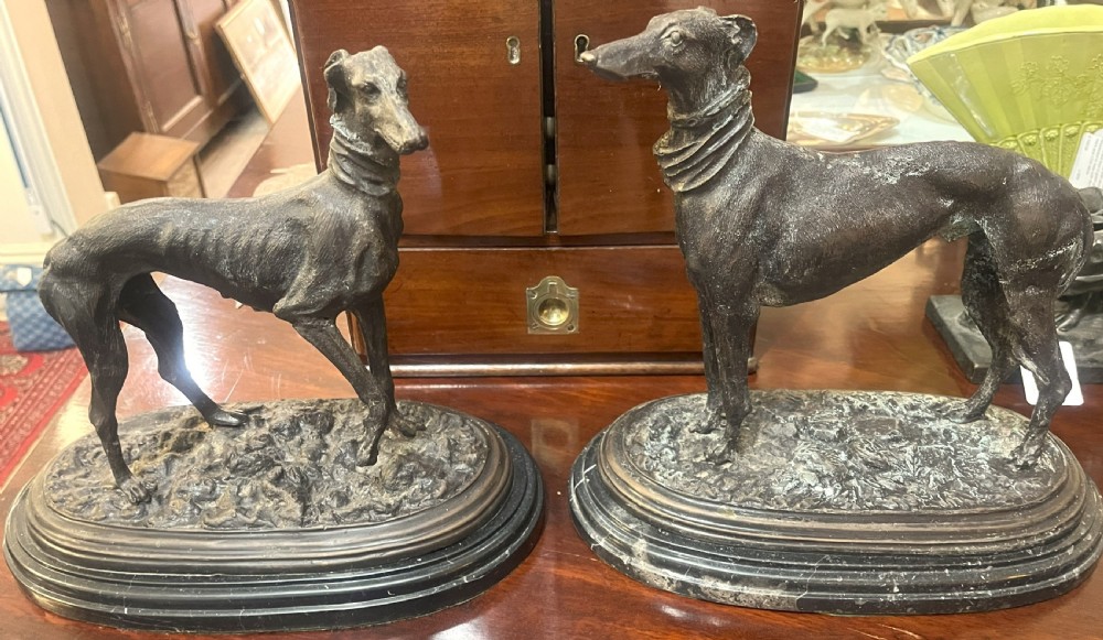A pair of patinated dark bronze whippet dog sculptures standing on molded oval black marble bases.