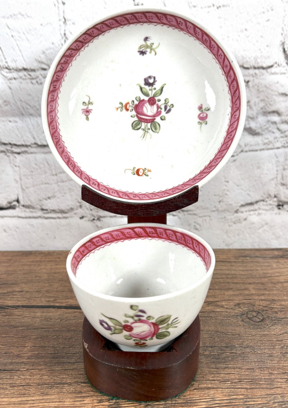 An antique white porcelain handleless tea bowl and matching saucer featuring hand-painted floral motifs and a pink ribbon border mounted on a dark wooden display stand.