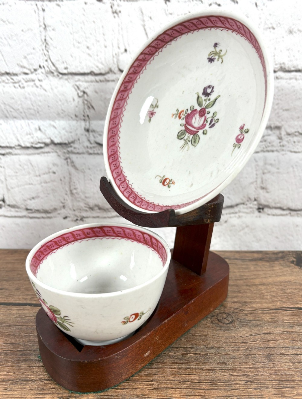 An antique white porcelain handleless tea bowl and matching saucer featuring hand-painted floral motifs and a pink ribbon border mounted on a dark wooden display stand. View II