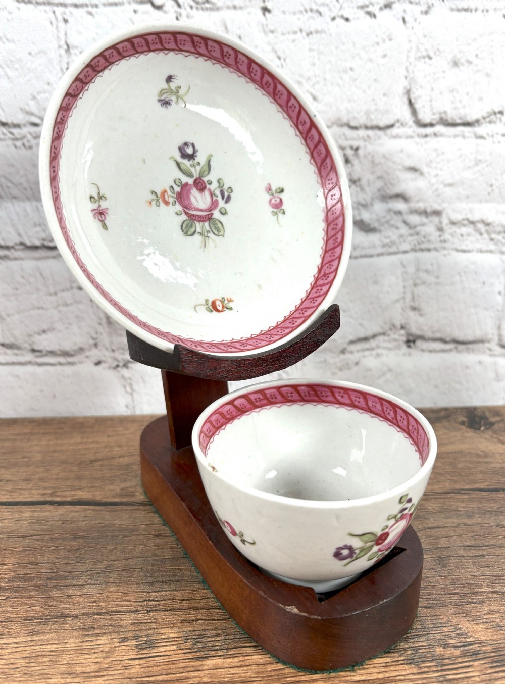 An antique white porcelain handleless tea bowl and matching saucer featuring hand-painted floral motifs and a pink ribbon border mounted on a dark wooden display stand. View III
