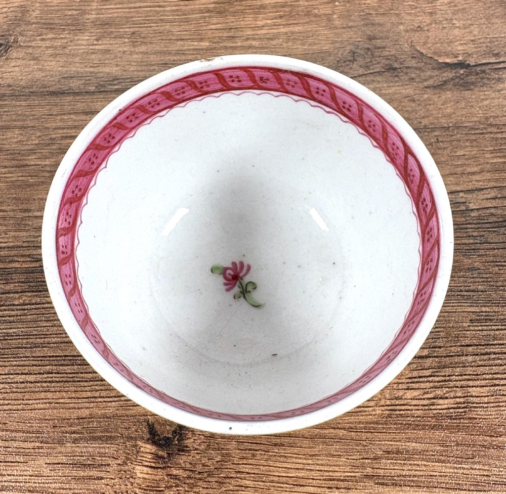 An antique white porcelain handleless tea bowl and matching saucer featuring hand-painted floral motifs and a pink ribbon border mounted on a dark wooden display stand. View IV
