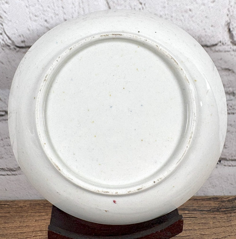 An antique white porcelain handleless tea bowl and matching saucer featuring hand-painted floral motifs and a pink ribbon border mounted on a dark wooden display stand. View V