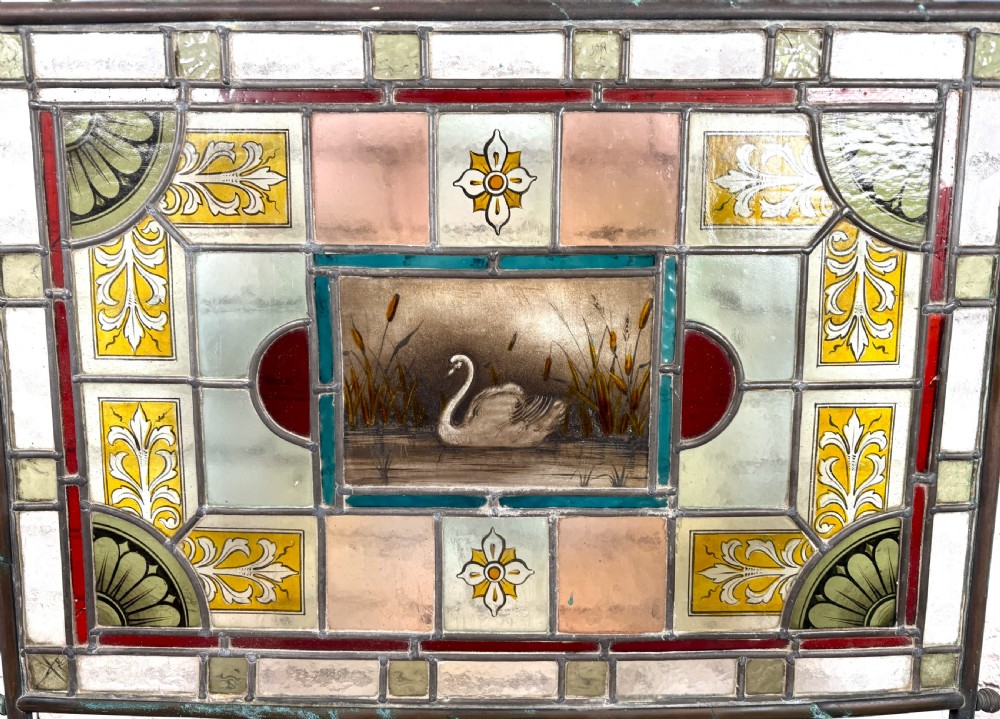 Victorian brass-framed fireguard featuring leaded stained glass panels in yellow, red, and clear geometric patterns surrounding a central hand-painted panel of a swan. View II
