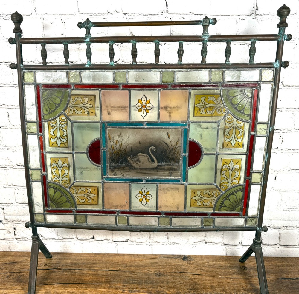 Victorian brass-framed fireguard featuring leaded stained glass panels in yellow, red, and clear geometric patterns surrounding a central hand-painted panel of a swan. View IV