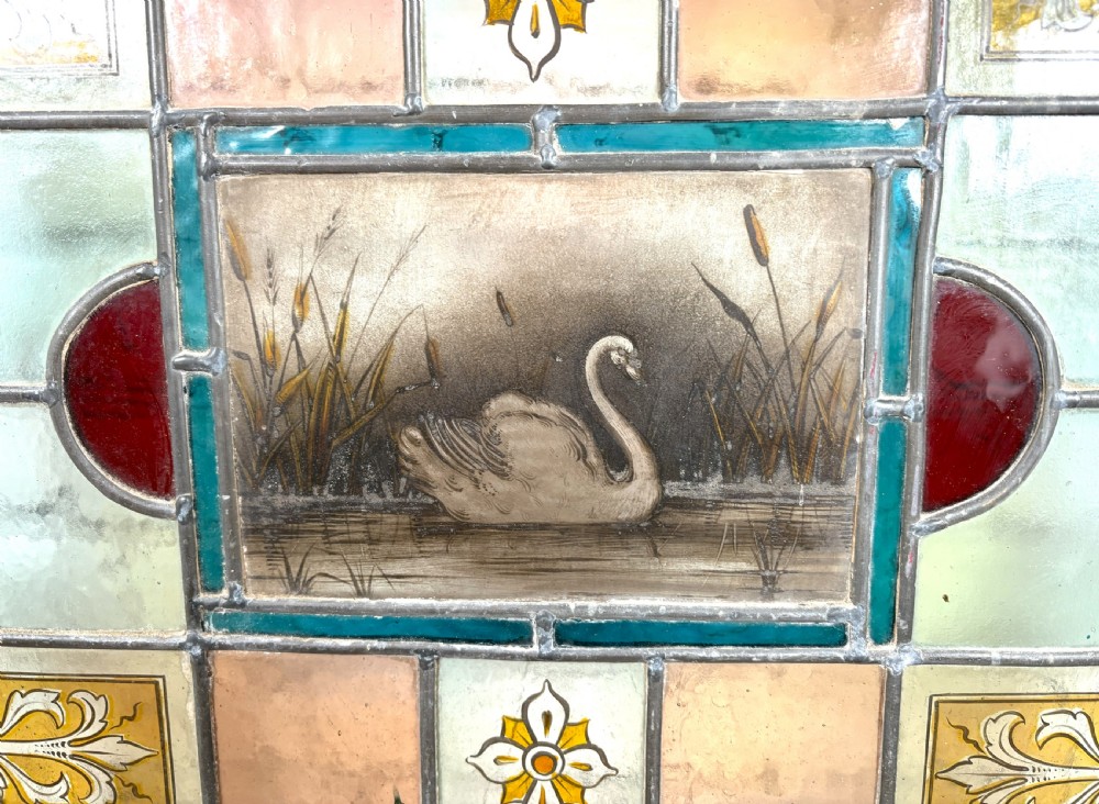 Victorian brass-framed fireguard featuring leaded stained glass panels in yellow, red, and clear geometric patterns surrounding a central hand-painted panel of a swan. View V