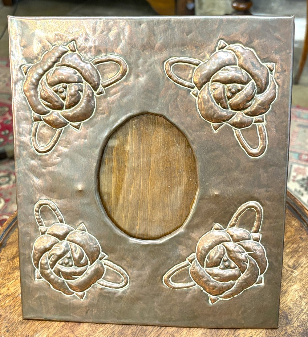 A hand-hammered rectangular copper picture frame featuring raised repousse rose motifs at each corner surrounding a central oval picture opening.