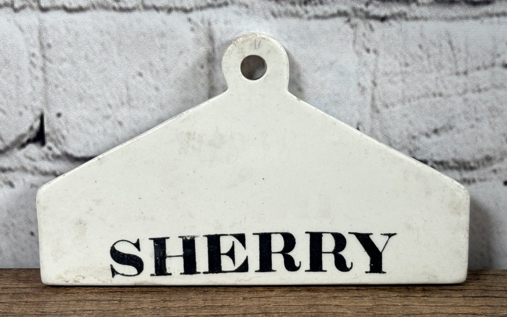 An antique off-white pottery wine bin label with a triangular top, hanging hole, and the word SHERRY printed in black serif text.