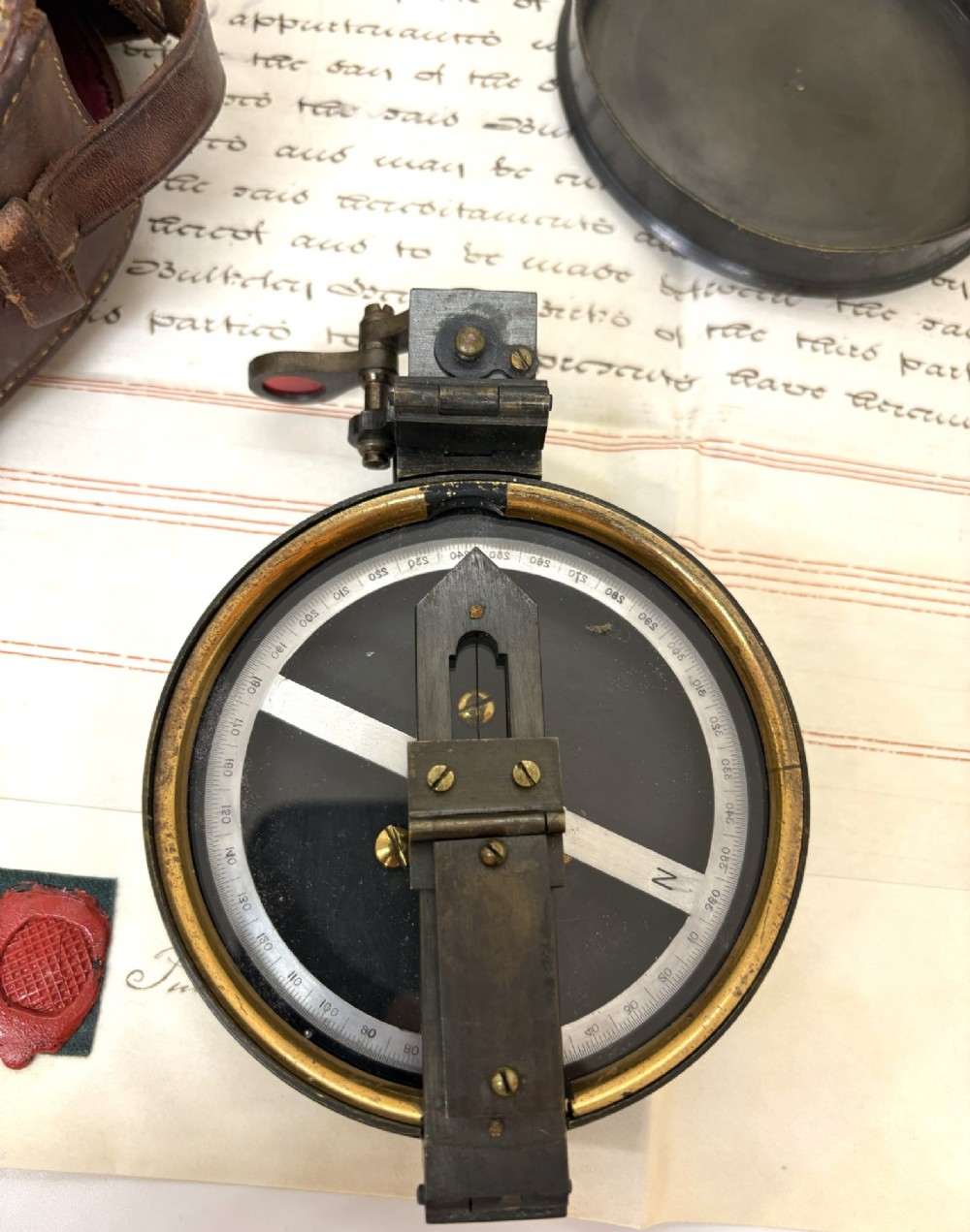 An early 20th-century blackened brass prismatic compass displayed open beside its original fitted brown leather carrying case. View II