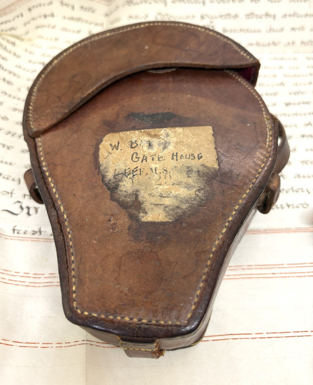 An early 20th-century blackened brass prismatic compass displayed open beside its original fitted brown leather carrying case. View III