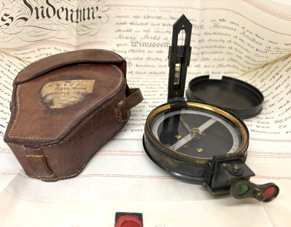 An early 20th-century blackened brass prismatic compass displayed open beside its original fitted brown leather carrying case. View IV