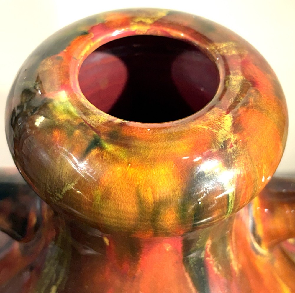 A bulbous ceramic vase featuring twin loop handles and a glossy multi-coloured running glaze in shades of red, green, and dark brown. View IV