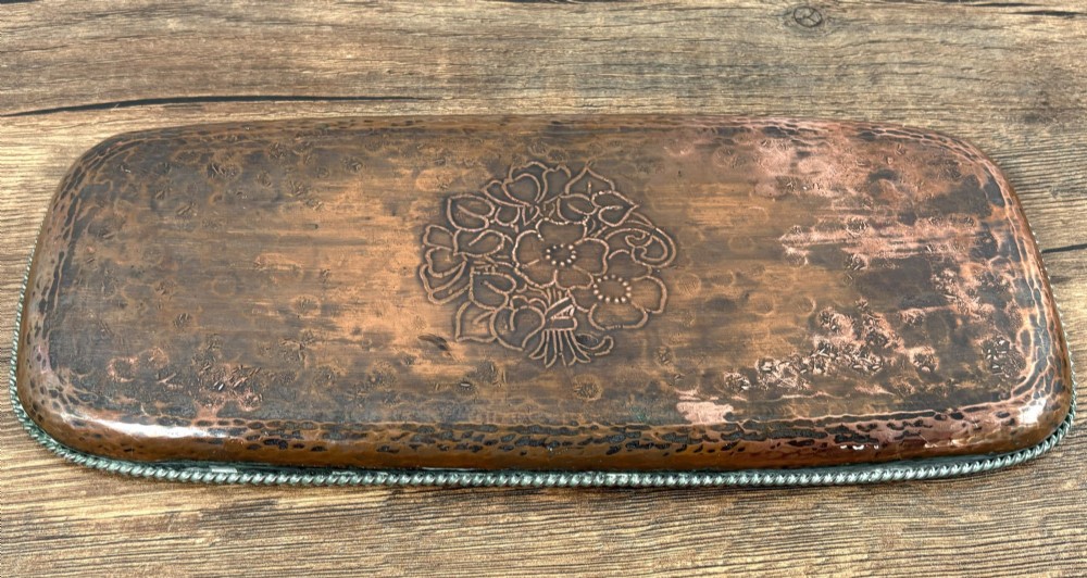 A rectangular hand-hammered copper tray featuring a central engraved floral bouquet motif, a rope-twist pewter rim, and a stamped makers mark signature. View V