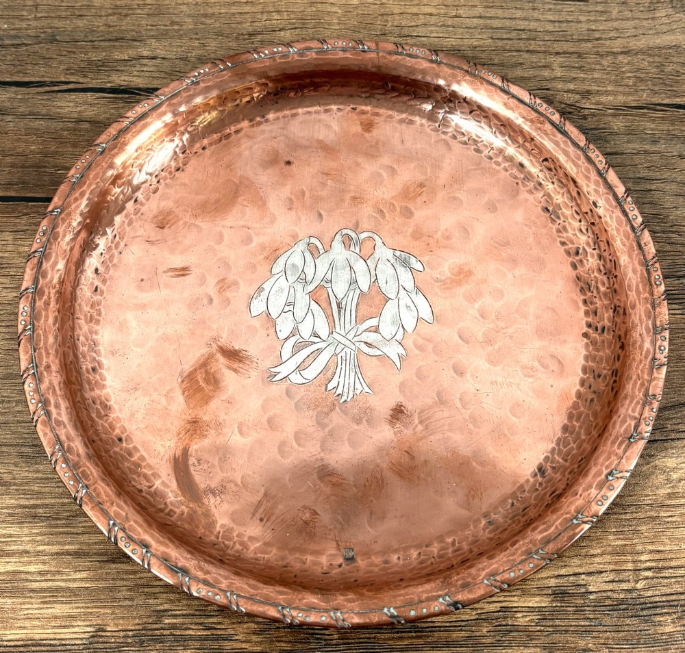 A circular hand-hammered copper tray featuring a pewter inlay of snowdrop flowers tied with a ribbon at the centre.