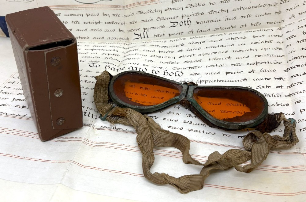 A pair of early 20th-century aviation goggles with amber glass lenses and a fabric strap resting alongside a brown storage case.