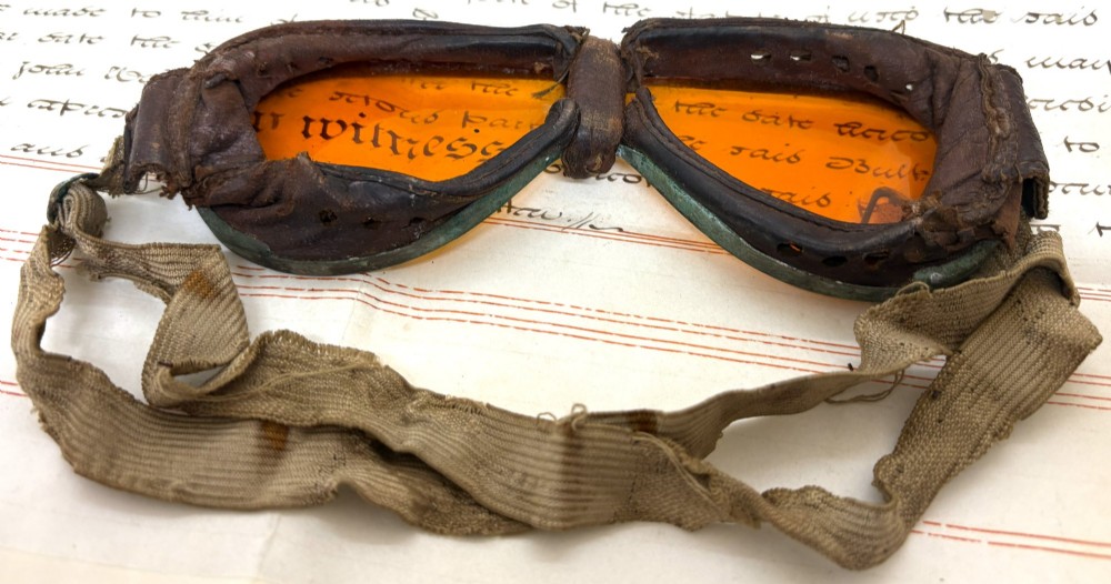 A pair of early 20th-century aviation goggles with amber glass lenses and a fabric strap resting alongside a brown storage case. View III