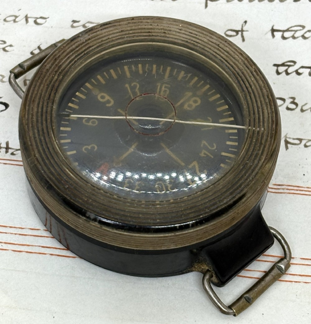 A circular black bakelite military wrist compass featuring a clear rotating acrylic bezel, numeric markings on a dark dial, and side metal strap loops. View II