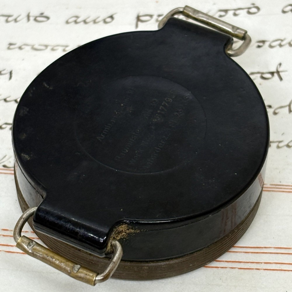 A circular black bakelite military wrist compass featuring a clear rotating acrylic bezel, numeric markings on a dark dial, and side metal strap loops. View III