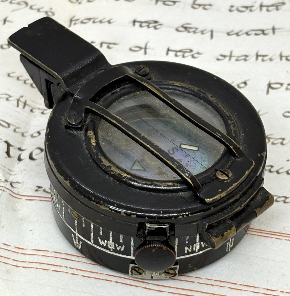 A circular black patinated brass military field compass featuring a central glass window, top sighting bar, and side directional markings. View II