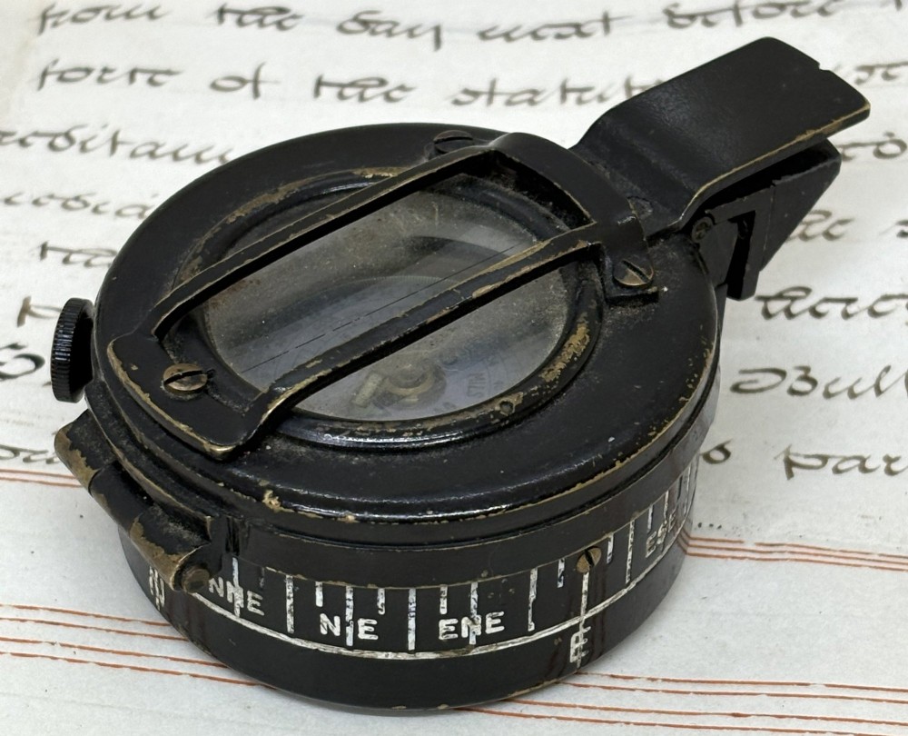 A circular black patinated brass military field compass featuring a central glass window, top sighting bar, and side directional markings. View III