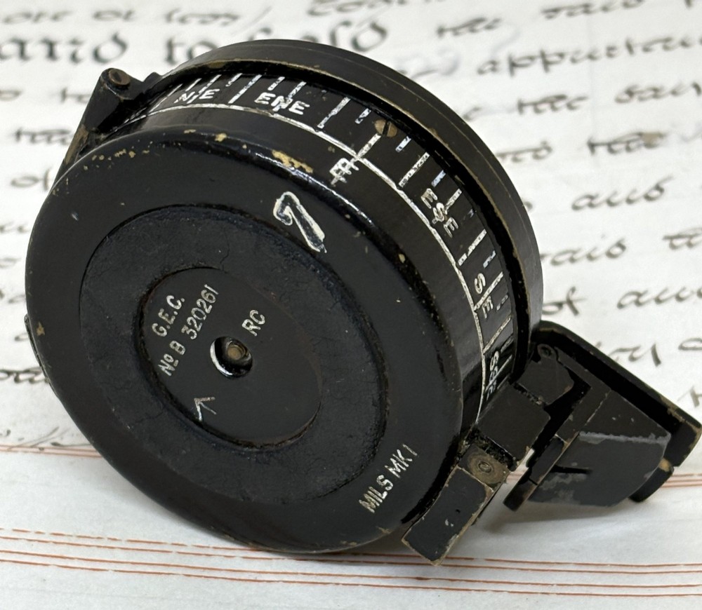 A circular black patinated brass military field compass featuring a central glass window, top sighting bar, and side directional markings. View IV