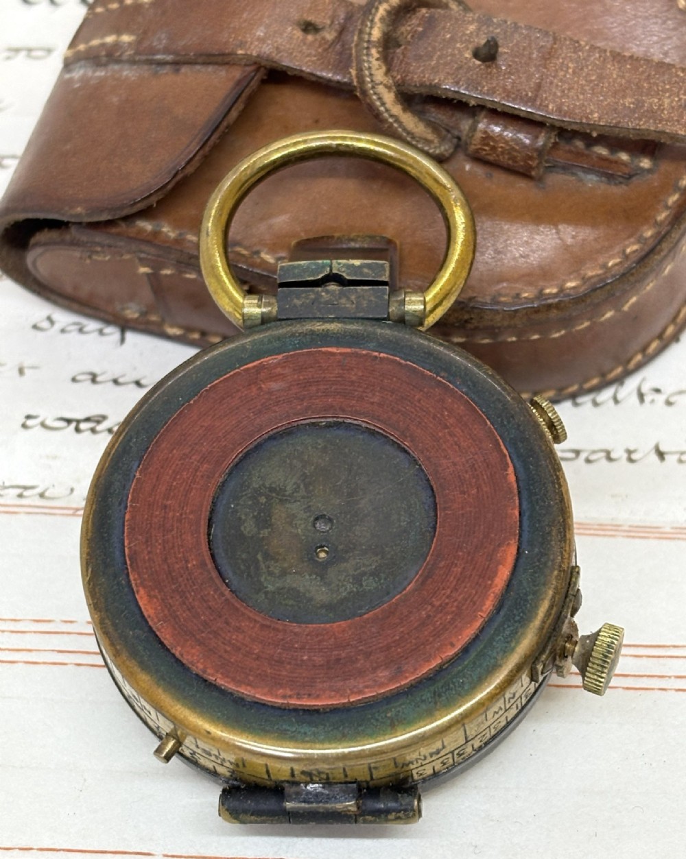 An antique brass pocket field compass resting on its original stitched brown leather protective case. View V