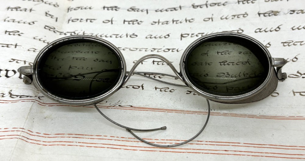 A pair of early twentieth-century metal goggles featuring dark tinted oval lenses, fine wire mesh side shields, and flexible cable arms.