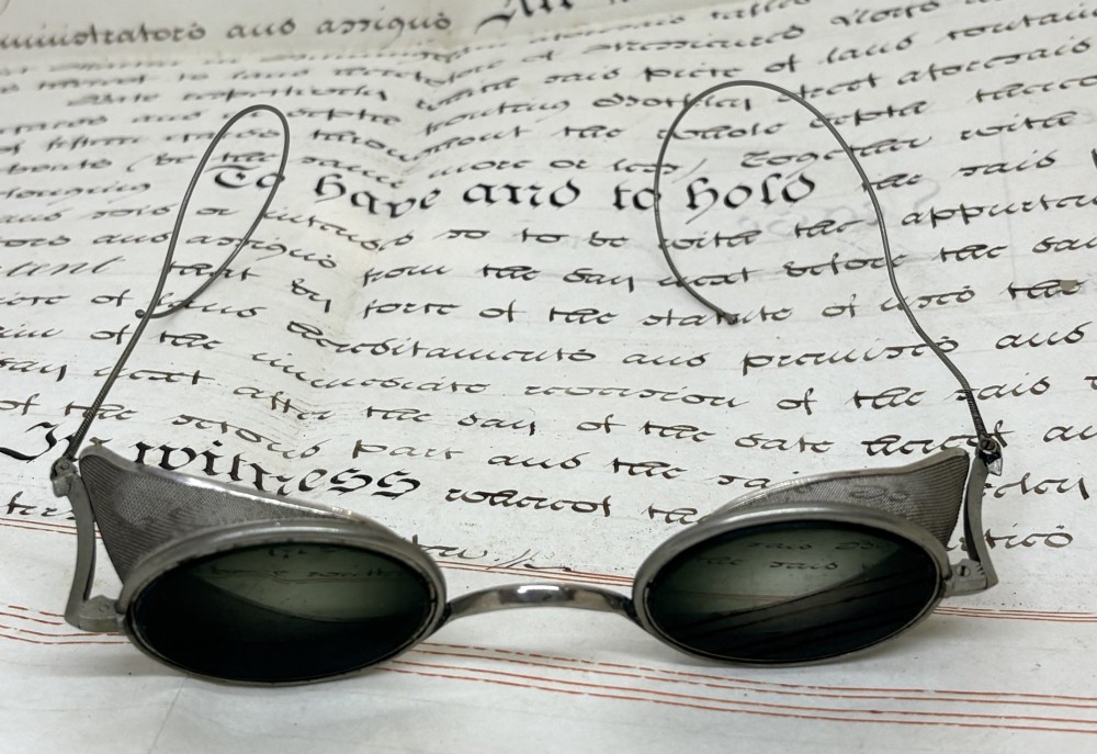 A pair of early twentieth-century metal goggles featuring dark tinted oval lenses, fine wire mesh side shields, and flexible cable arms. View II