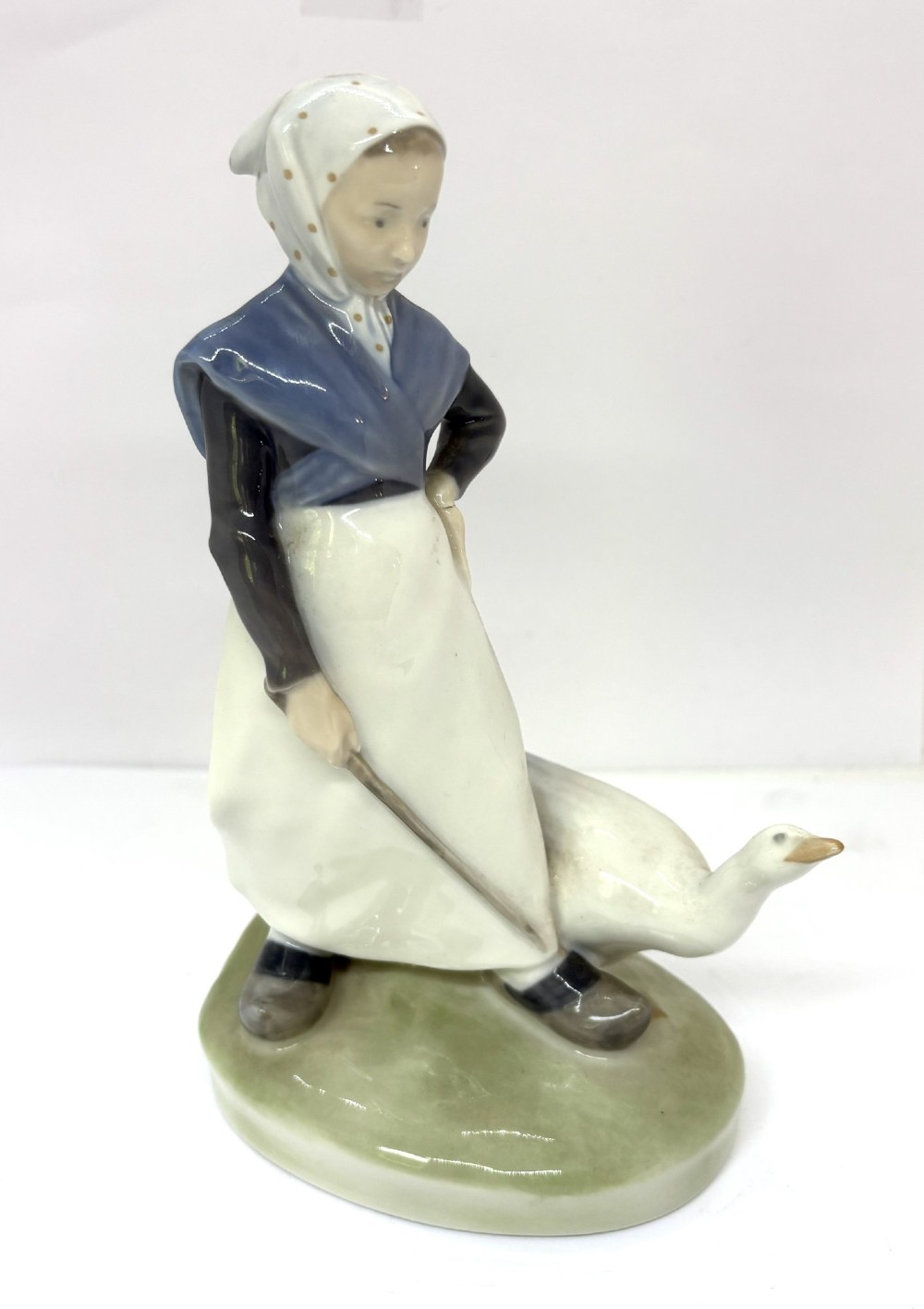 A glazed Royal Copenhagen porcelain figurine depicting a girl in a headscarf and shawl standing beside a white goose on an oval green base.