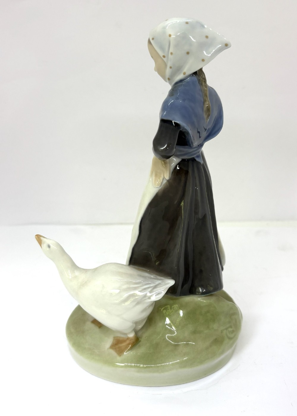 A glazed Royal Copenhagen porcelain figurine depicting a girl in a headscarf and shawl standing beside a white goose on an oval green base. View II