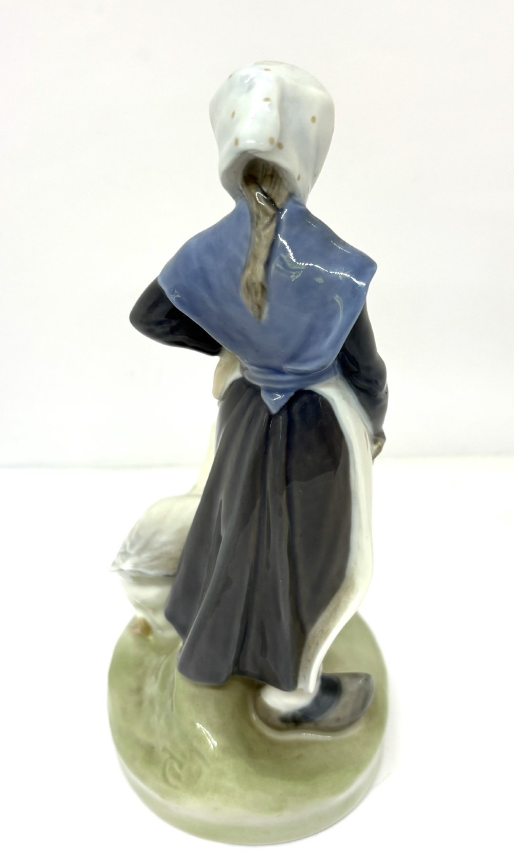 A glazed Royal Copenhagen porcelain figurine depicting a girl in a headscarf and shawl standing beside a white goose on an oval green base. View III