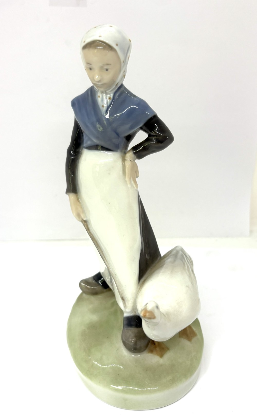 A glazed Royal Copenhagen porcelain figurine depicting a girl in a headscarf and shawl standing beside a white goose on an oval green base. View IV
