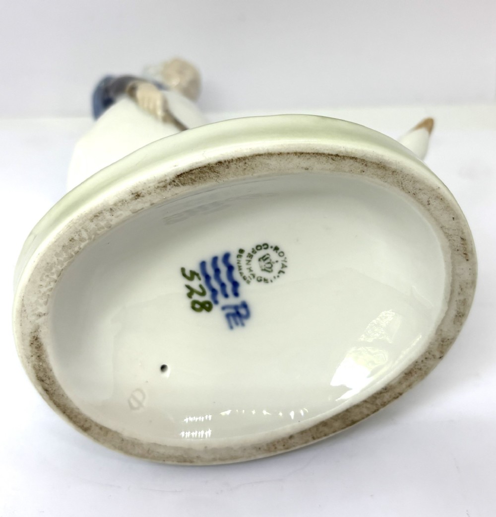 A glazed Royal Copenhagen porcelain figurine depicting a girl in a headscarf and shawl standing beside a white goose on an oval green base. View V