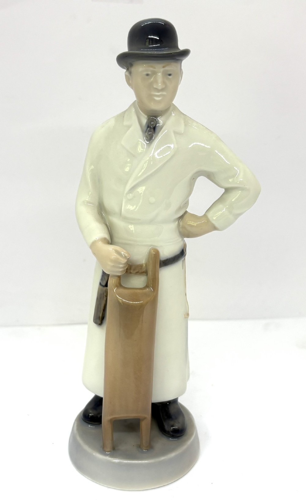A glazed porcelain figurine depicting a butcher wearing a bowler hat, double-breasted coat, and apron standing beside a brown trough.